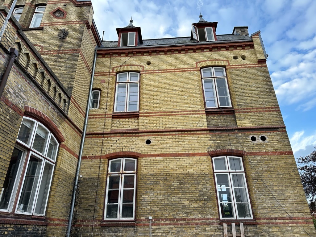 Żółta ceglana fasada budynku z czerwonymi akcentami, widoczne okna, fragment dachu z lukarnami i drabina oparta o ścianę.