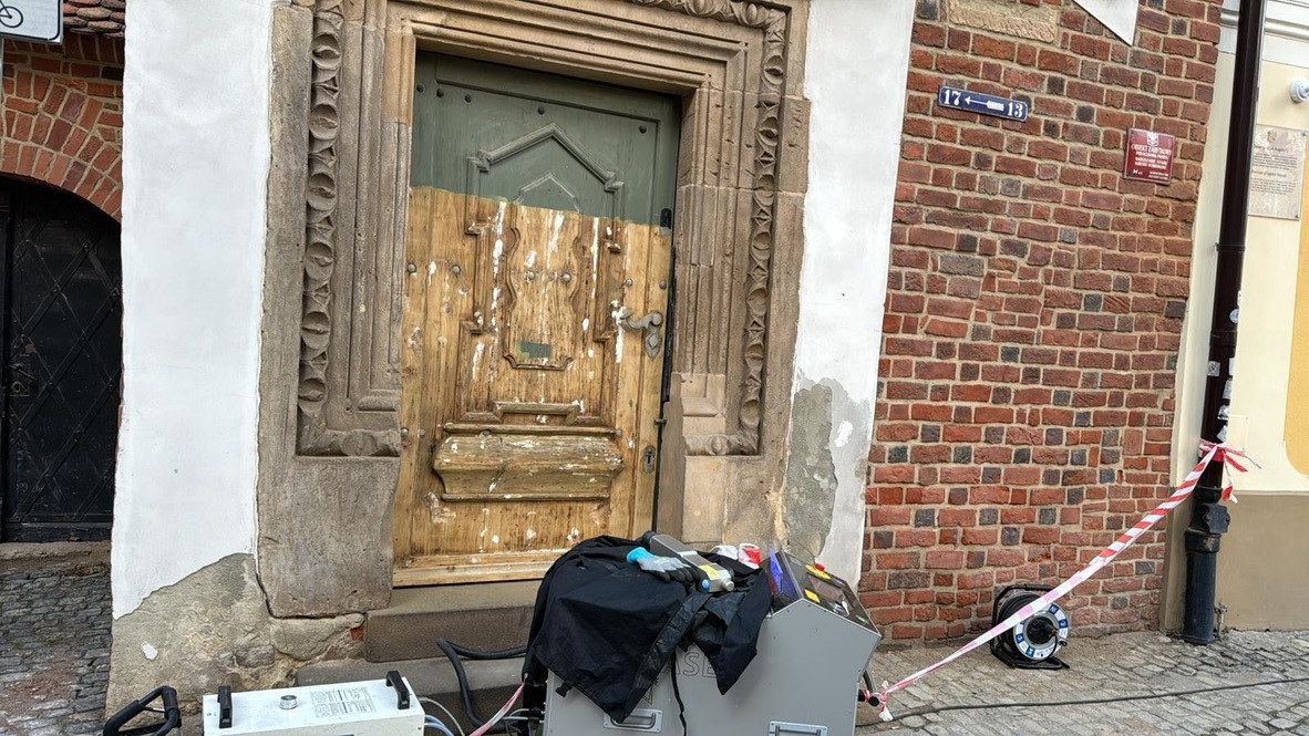 Stara drewniana brama z kamiennym obramowaniem w trakcie renowacji, z widocznym sprzętem do czyszczenia i zabezpieczającą taśmą.