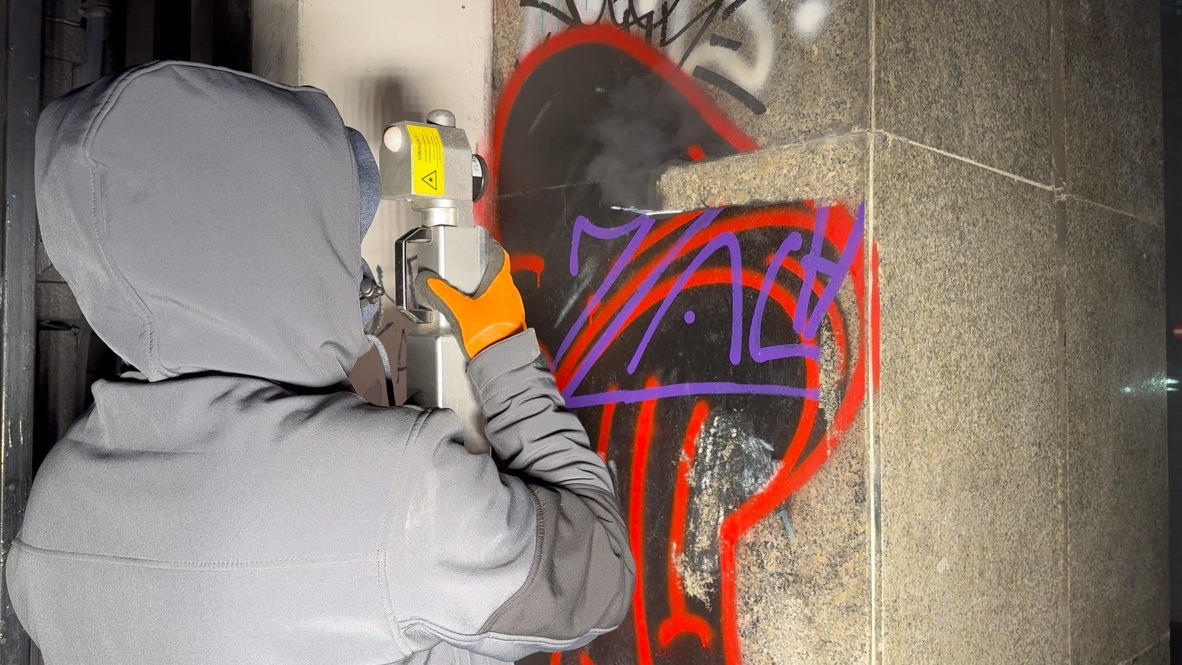 Osoba w szarej kurtce z kapturem i pomarańczowych rękawicach usuwa graffiti z kamiennej ściany za pomocą specjalistycznego urządzenia laserowego, z którego wydobywa się dym.