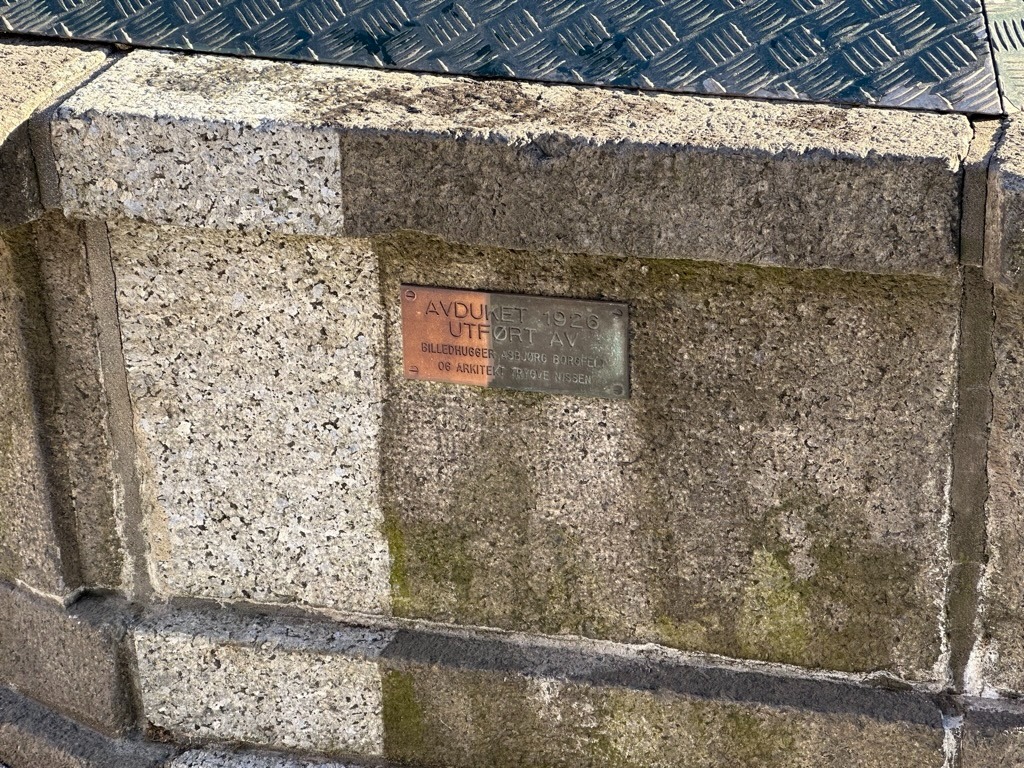 Fragment kamiennej konstrukcji z mosiężną tabliczką pamiątkową, częściowo porośnięty mchem, z widoczną metalową powierzchnią w tle.