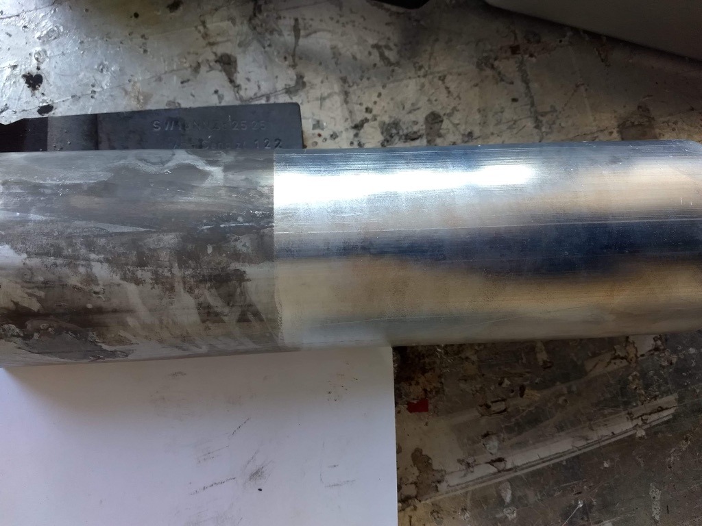 Metalowy element cylindryczny, w połowie oczyszczony z zabrudzeń, leżący na białym arkuszu papieru na tle betonowej podłogi z plamami.