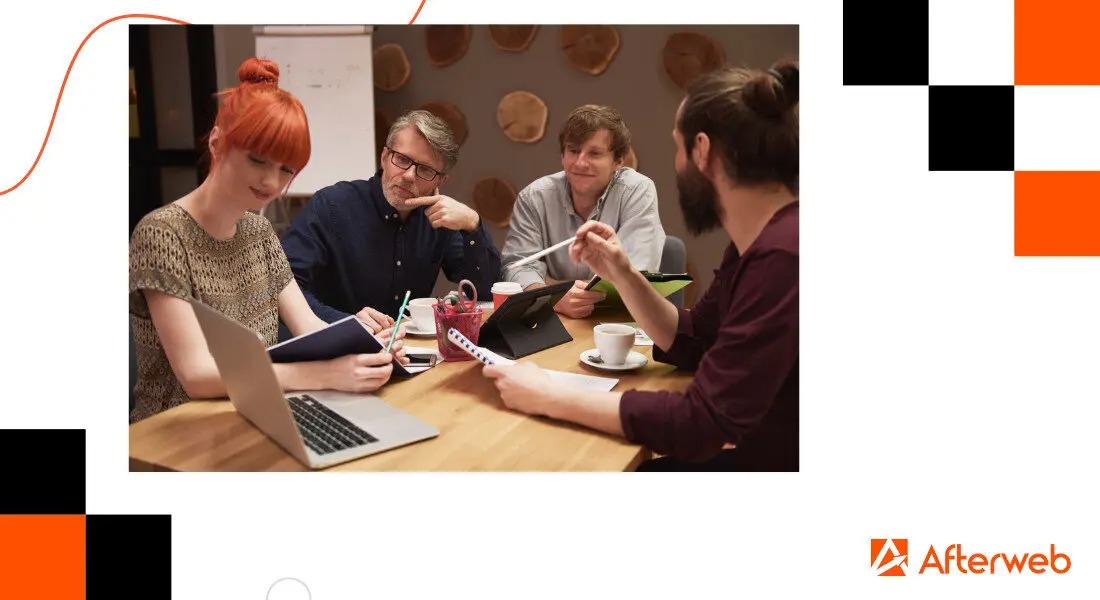 Zespół czterech osób przy jasnym drewnianym stole, omawiający strategię, z laptopem, notatkami, tabletem i kawą, w tle ściana z drewnianymi dekoracjami.