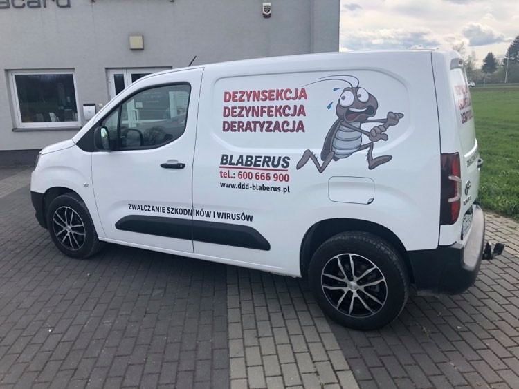 Biały samochód dostawczy z reklamą firmy zajmującej się zwalczaniem szkodników i wirusów, z dużym rysunkiem uciekającego karalucha na boku.