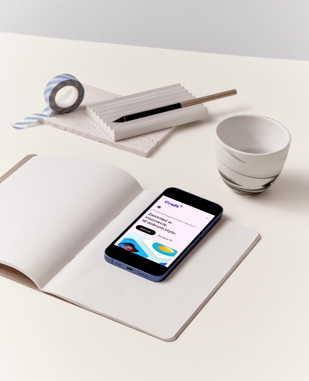 Smartfon wyświetlający stronę internetową na otwartym notatniku, obok kubek, taśma washi i pióro w ceramicznym uchwycie, minimalistyczna kompozycja na jasnym tle.