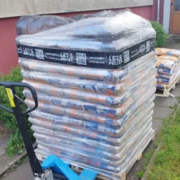 Stos worków z pelletem owiniętych folią na palecie, obok wózek paletowy, w tle budynek.