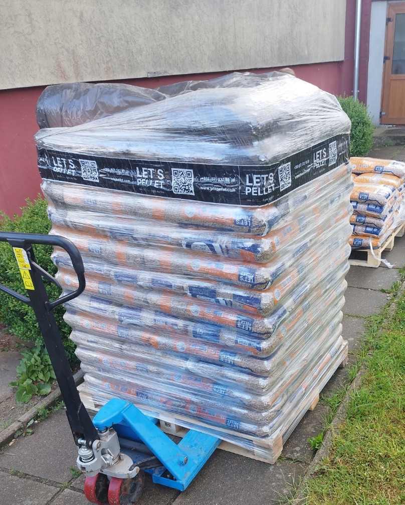 Stos worków z pelletem owiniętych folią na palecie, obok wózek paletowy, w tle budynek.