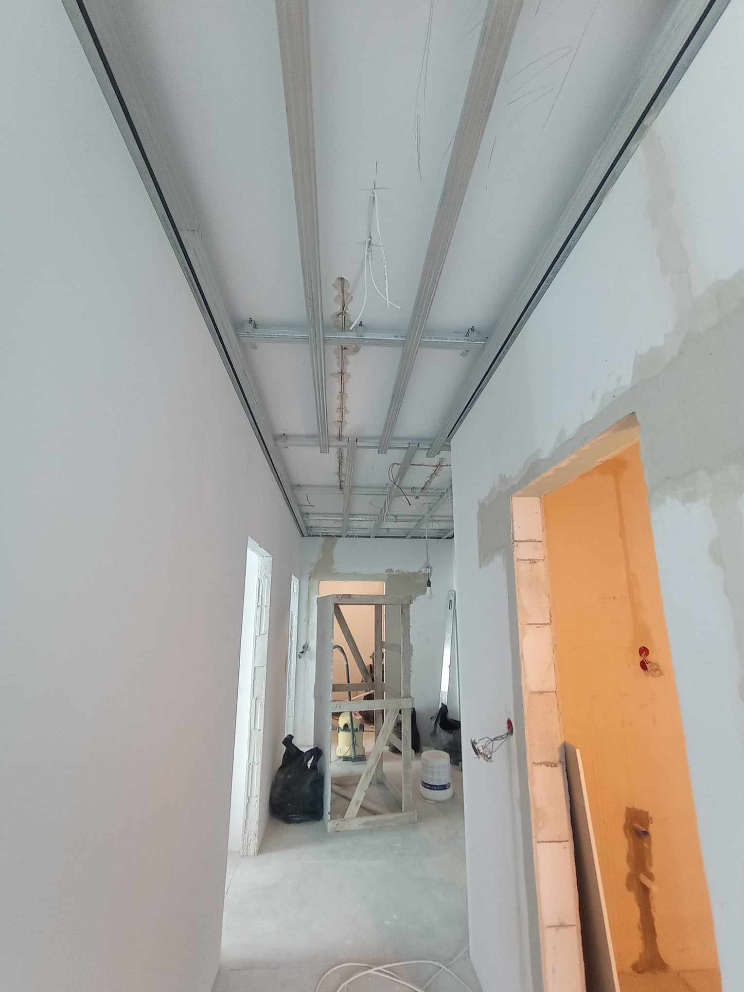 Montaż sufitu podwieszanego z widoczną konstrukcją metalową, przewody elektryczne wystające z sufitu, ściany w trakcie szpachlowania, worek z odpadami i rusztowanie w tle.