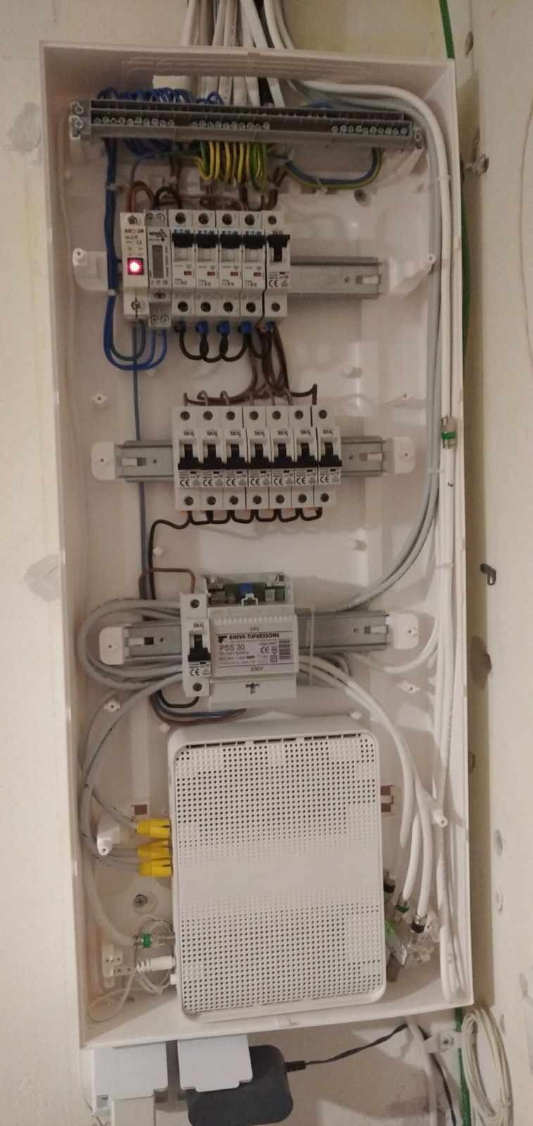 Otwarta skrzynka elektryczna z zainstalowanymi bezpiecznikami, przewodami, transformatorem i routerem światłowodowym z podłączonymi kablami Ethernet w kolorze żółtym.