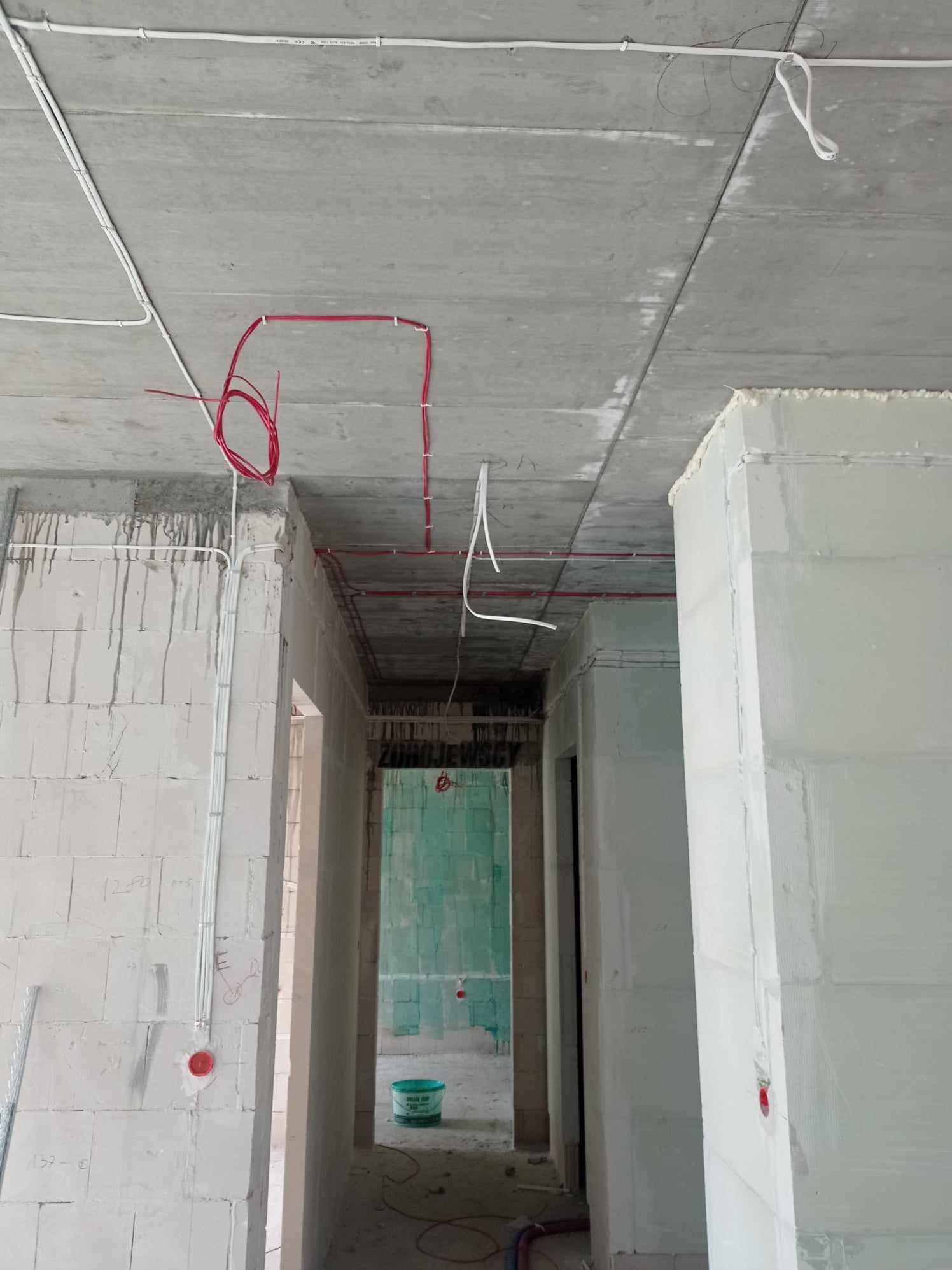 Surowy stan budynku w trakcie instalacji elektrycznej: widoczne kable w kolorach białym i czerwonym, prowadzone po betonowym suficie i ścianach z bloczków, z puszkami instalacyjnymi na ścianach,...