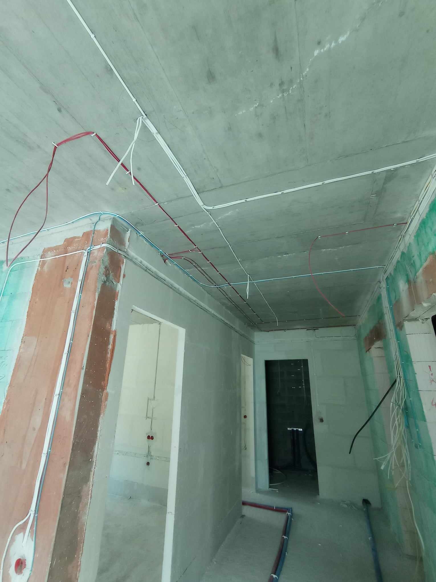 Surowy stan wnętrza w trakcie prac instalacyjnych: widoczne kable elektryczne prowadzone po suficie i ścianach, częściowo w korytach, w kolorach czerwonym, niebieskim i białym, surowy betonowy...