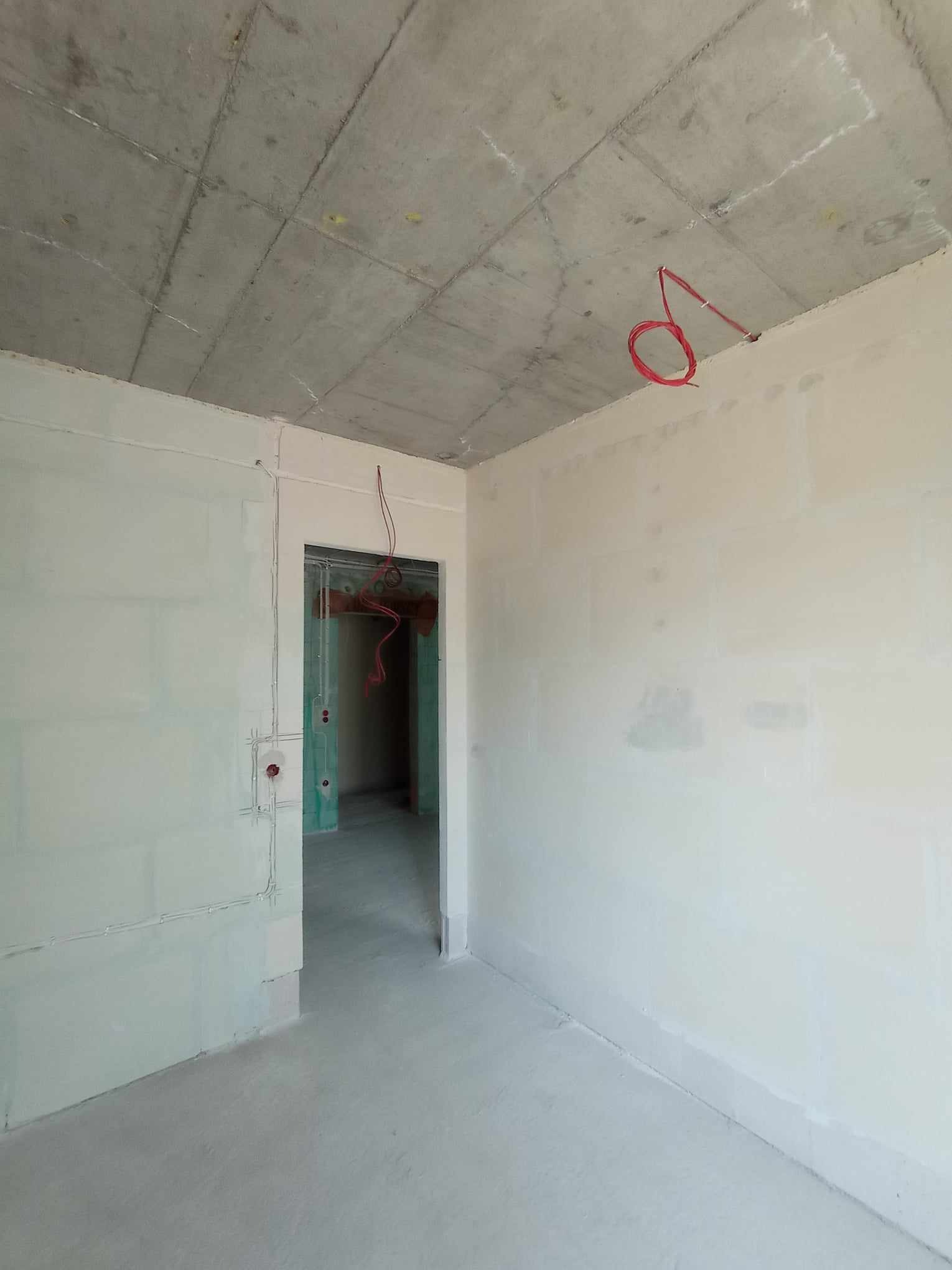 Surowy stan wnętrza z widocznymi przewodami elektrycznymi wystającymi ze ściany i sufitu, instalacja w trakcie realizacji, betonowy sufit i gładkie ściany.