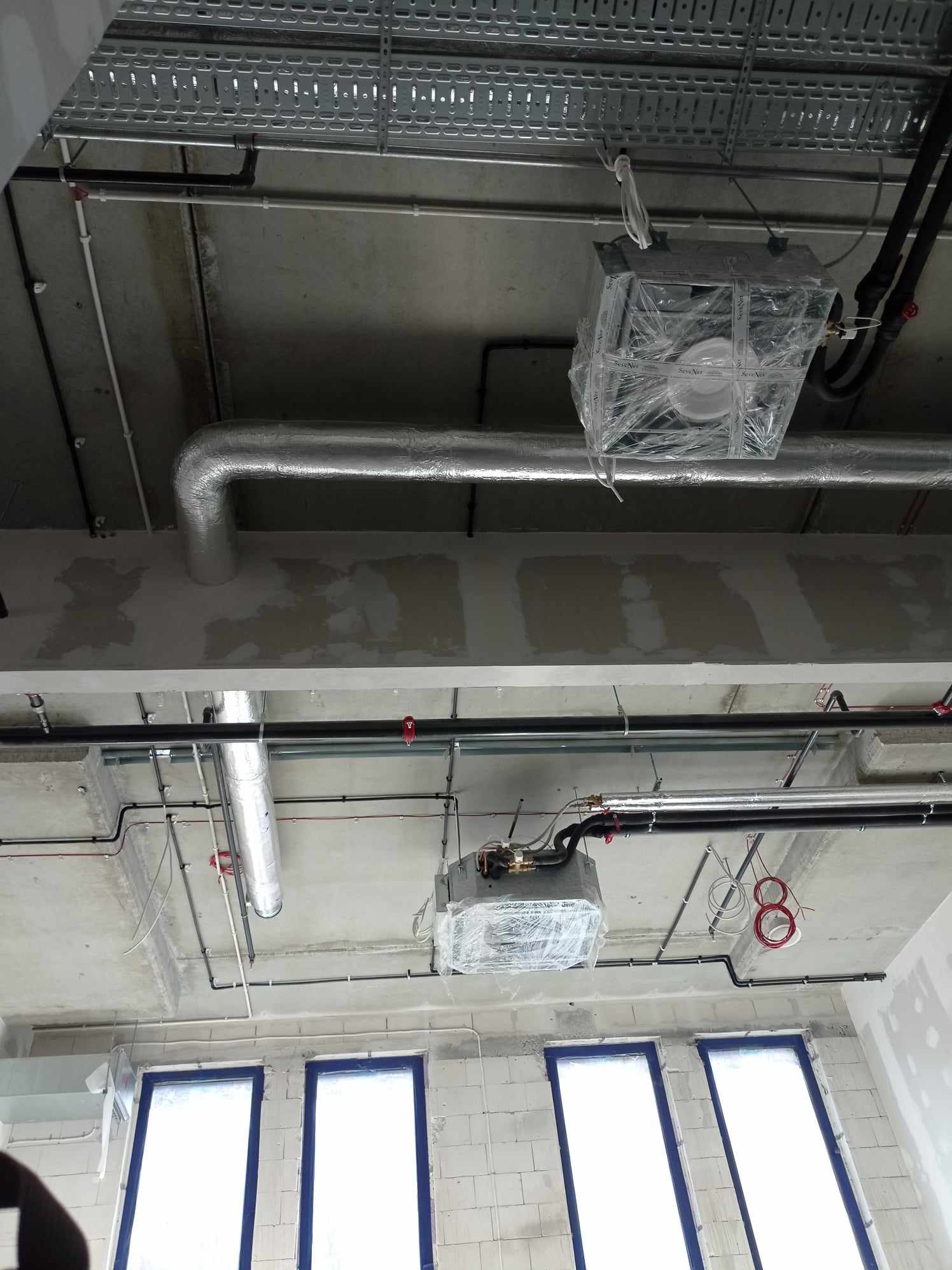 Widok sufitu w trakcie instalacji elektrycznej i wentylacyjnej, z widocznymi przewodami, rurami wentylacyjnymi w izolacji, korytkami kablowymi i lampami zabezpieczonymi folią, surowy beton i okna...