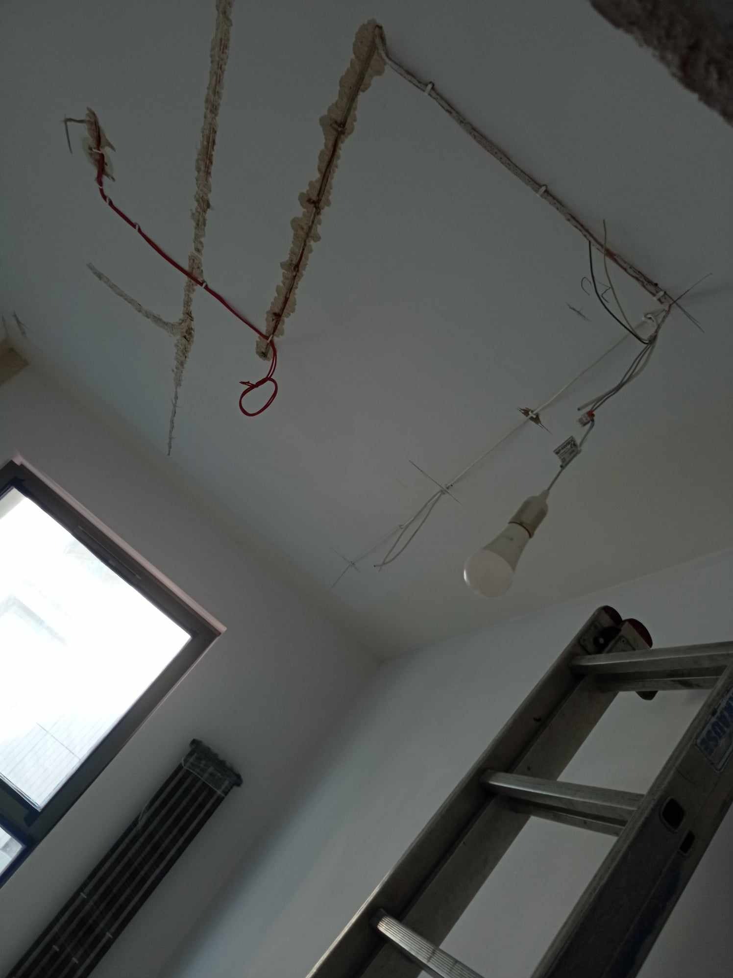 Niedokończona instalacja elektryczna w suficie z widocznymi przewodami, pianką montażową, żarówką na kablu i fragmentem drabiny.