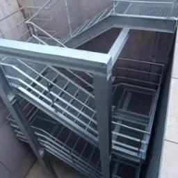 Metalowe schody z kratką w betonowym szybie, widok z góry, oświetlenie naturalne.