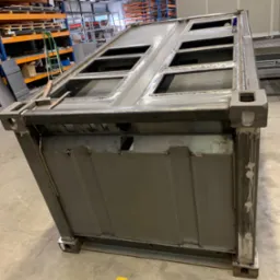 Częściowo zespawana metalowa konstrukcja skrzyni z otworami, widoczne spawy i ślady obróbki, na tle hali produkcyjnej z regałami.