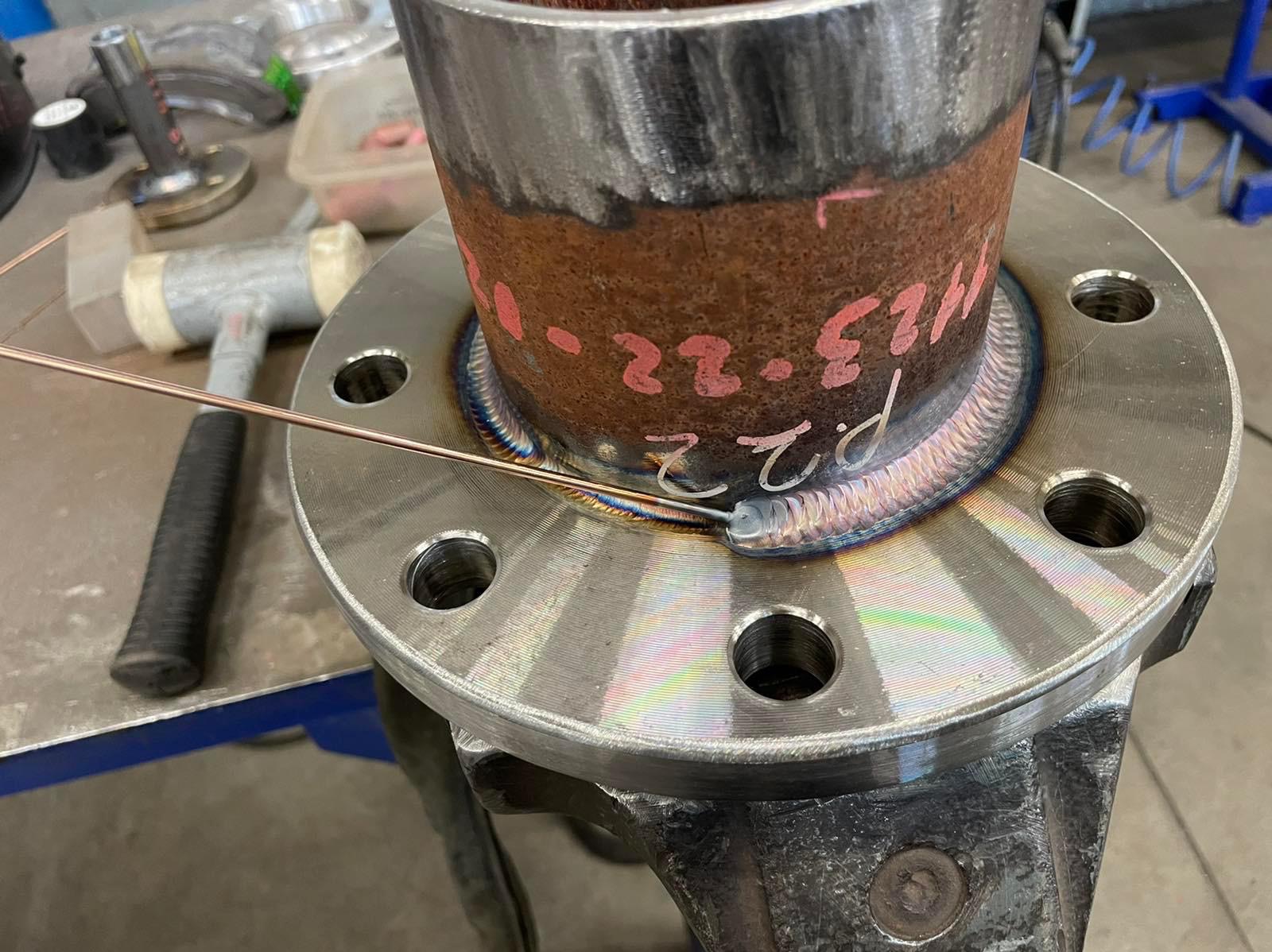 Fragment spawania rury do kołnierza stalowego, widoczny drut spawalniczy, świeży spaw z tęczowymi przebarwieniami, notatki na rurze, młotek gumowy w tle.