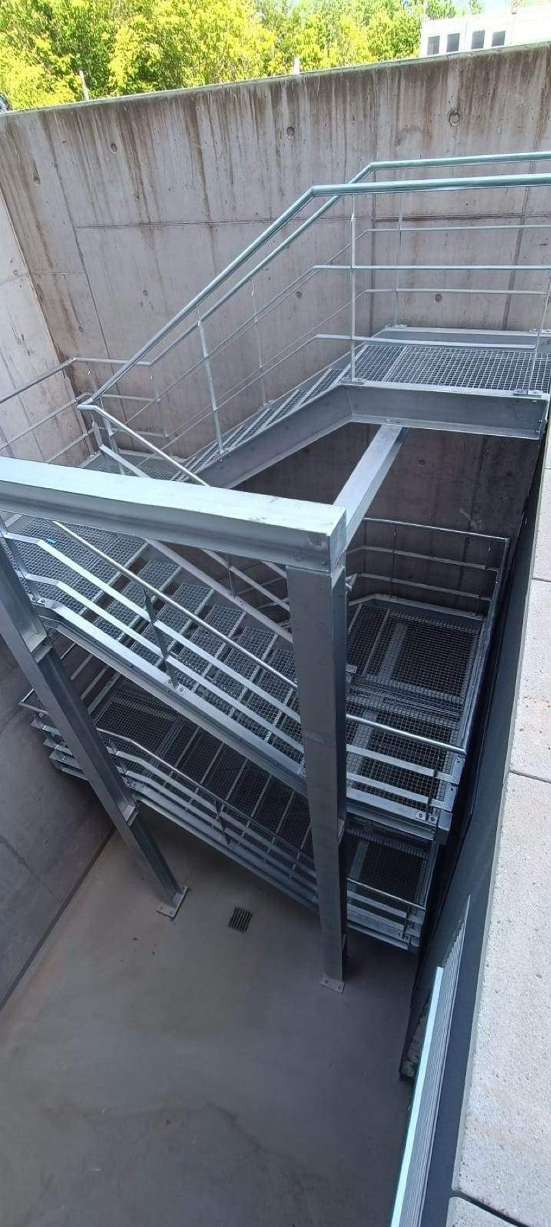 Metalowe schody z kratką w betonowym szybie, widok z góry, oświetlenie naturalne.