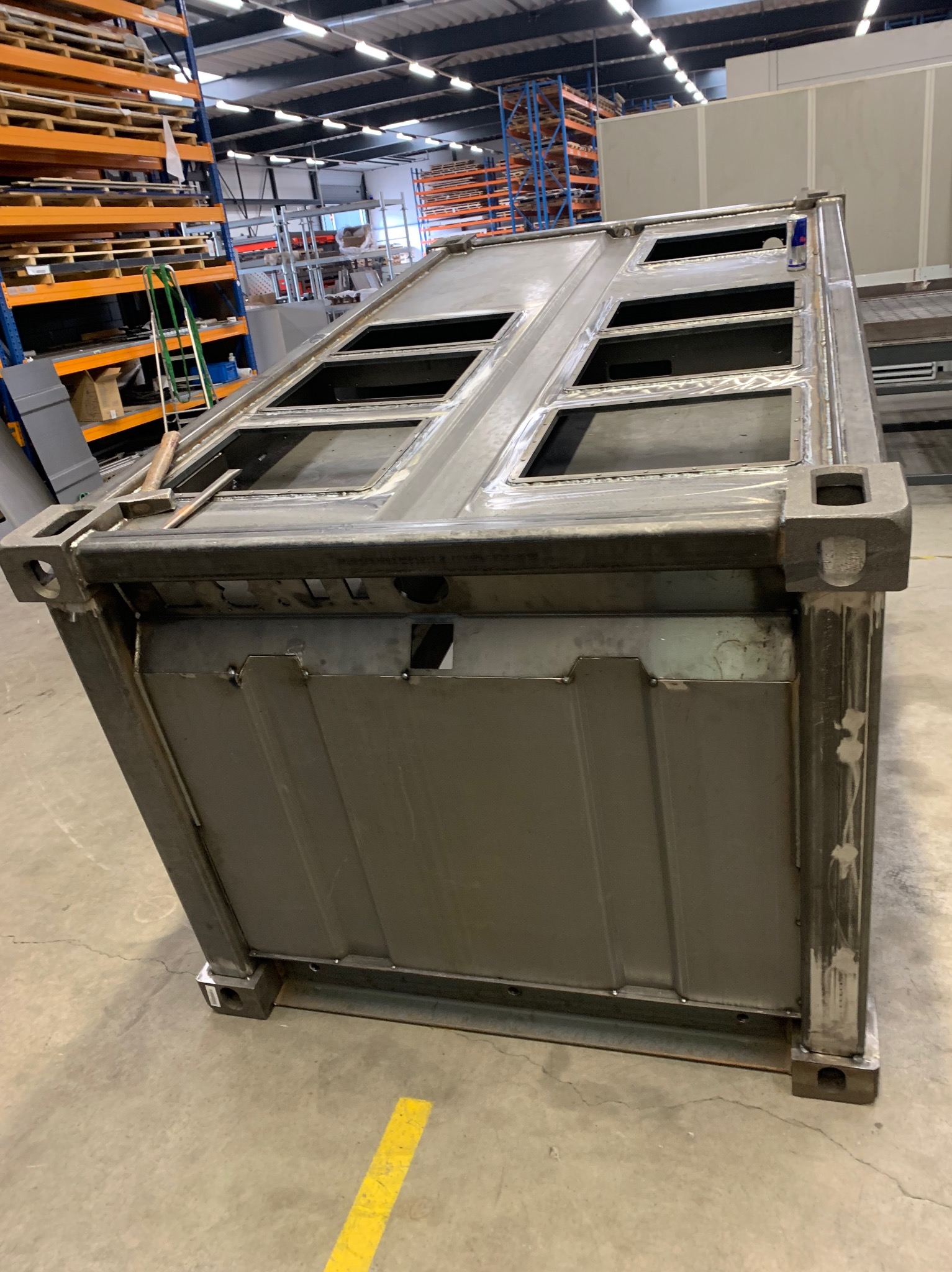 Częściowo zespawana metalowa konstrukcja skrzyni z otworami, widoczne spawy i ślady obróbki, na tle hali produkcyjnej z regałami.