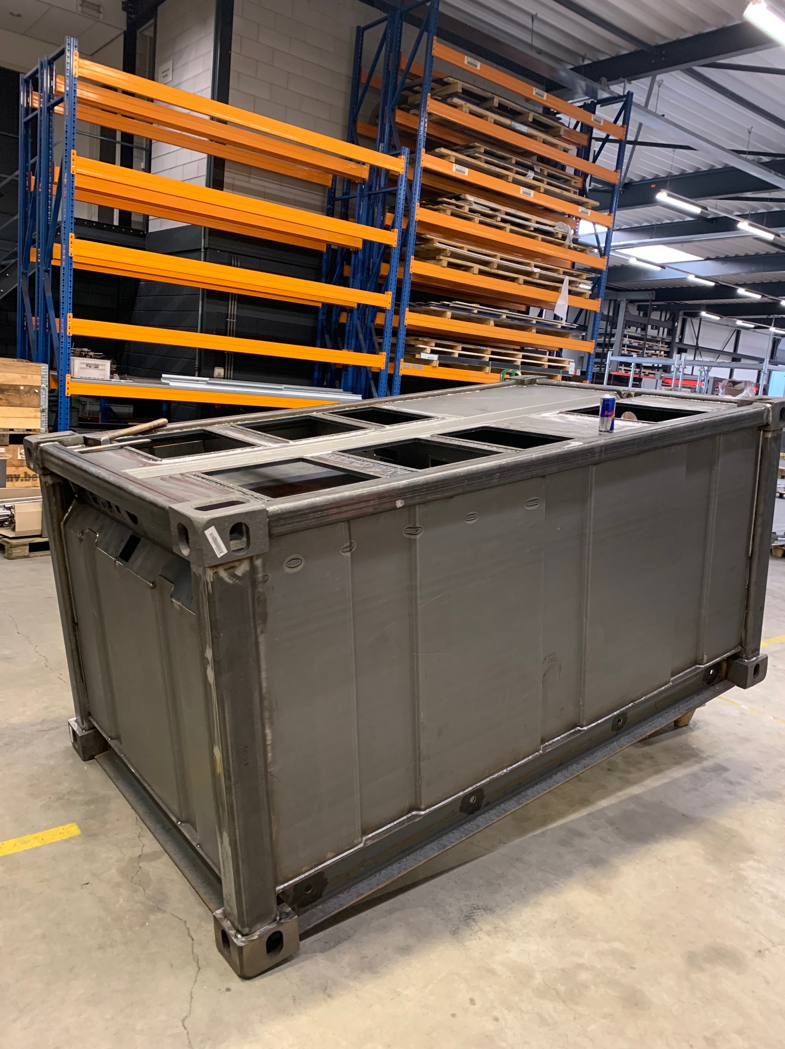 Szara, metalowa konstrukcja skrzyniowa z otworami w górnej części, widoczna w hali produkcyjnej z regałami w tle.