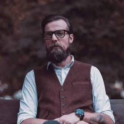 Paulina Wyszyńska-Górecka Photography & Marketing - Portret brodatego mężczyzny w okularach, brązowej kamizelce i białej koszuli, siedzącego na ławce z zamyślonym wyrazem twarzy.