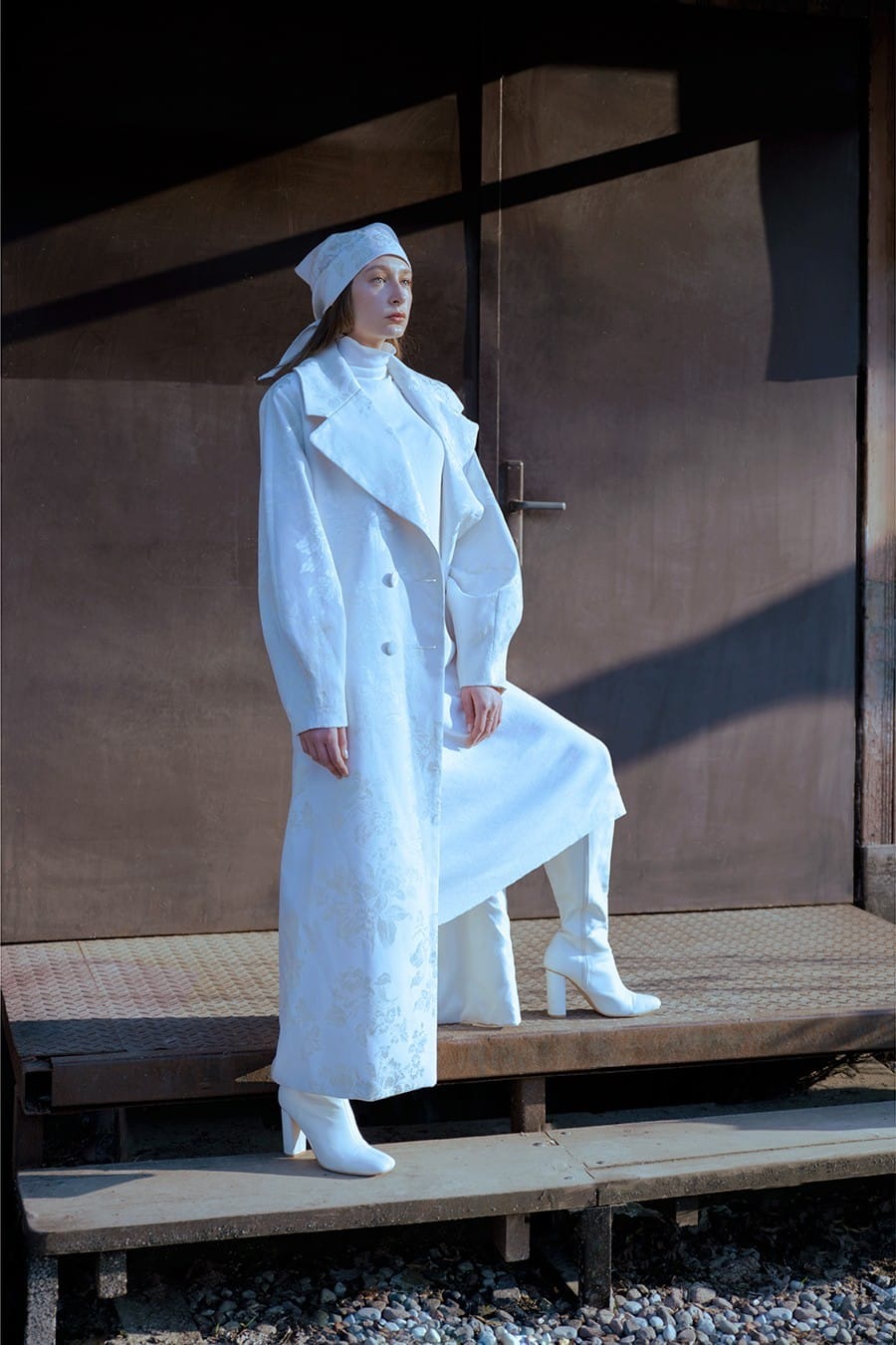 Kobieta w białym, wzorzystym płaszczu i kozakach na tle metalowych drzwi z cieniem.