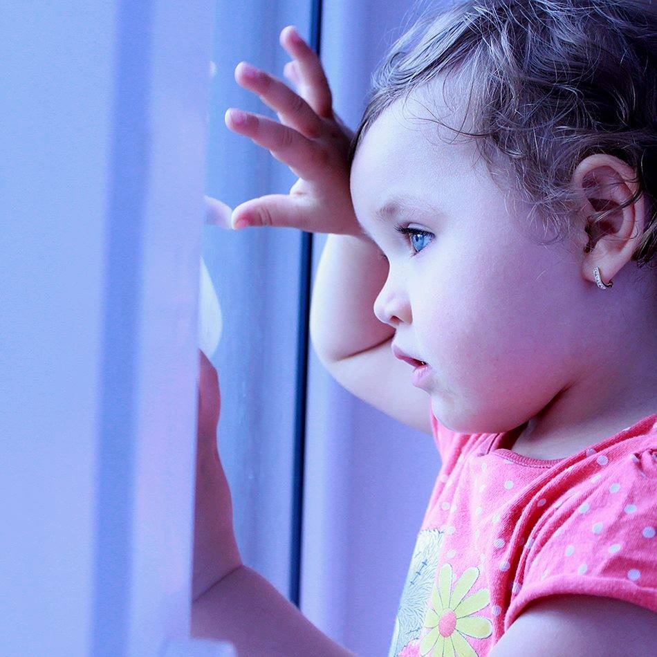 Dziewczynka z niebieskimi oczami, w różowej koszulce w kropki, opiera dłoń o szybę okna, patrząc na zewnątrz.