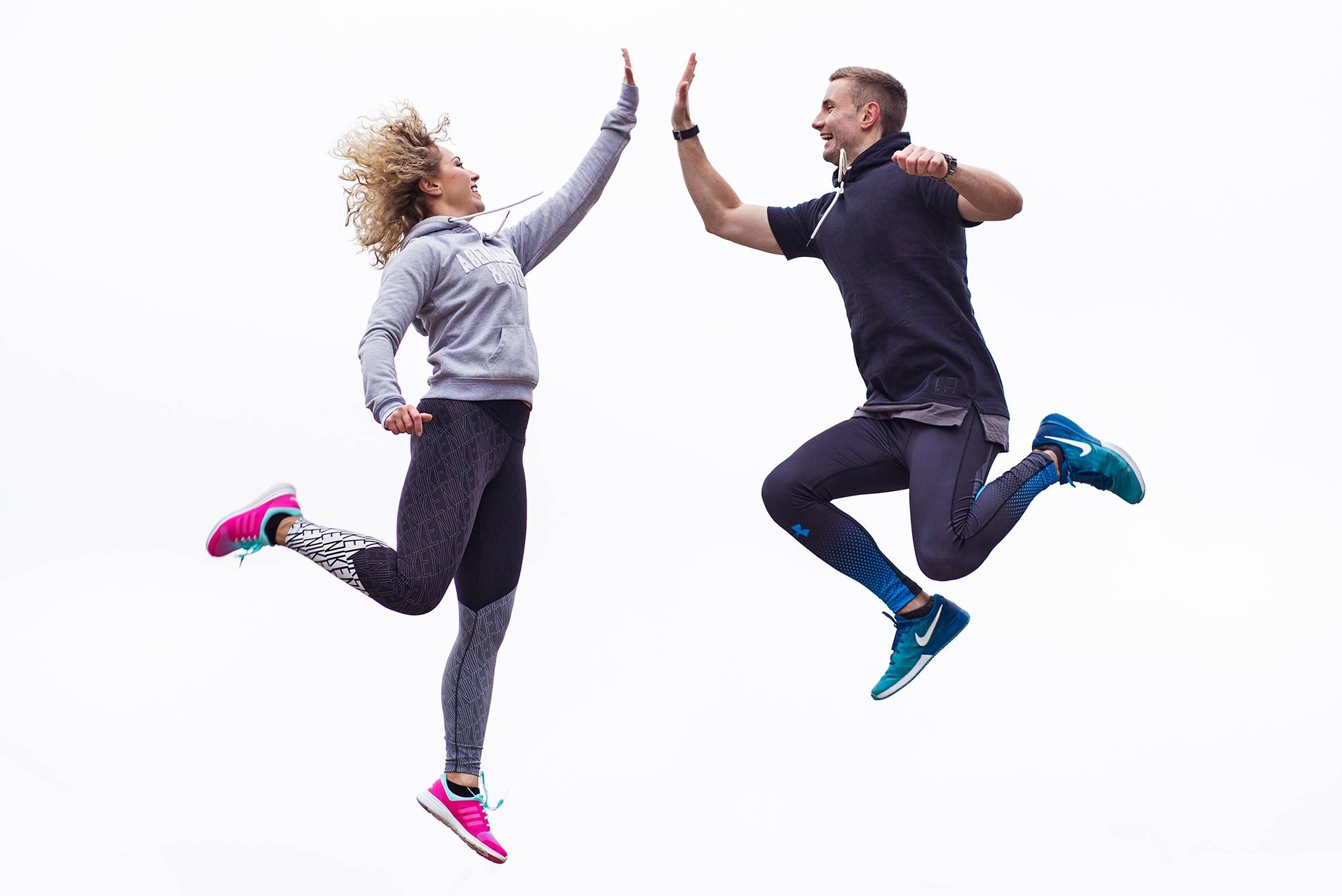 Dwoje sportowców w skoku, dający sobie piątkę na tle jasnego nieba, ona w szarej bluzie i wzorzystych legginsach, on w ciemnej koszulce i legginsach z gradientem, oboje w sportowych butach.