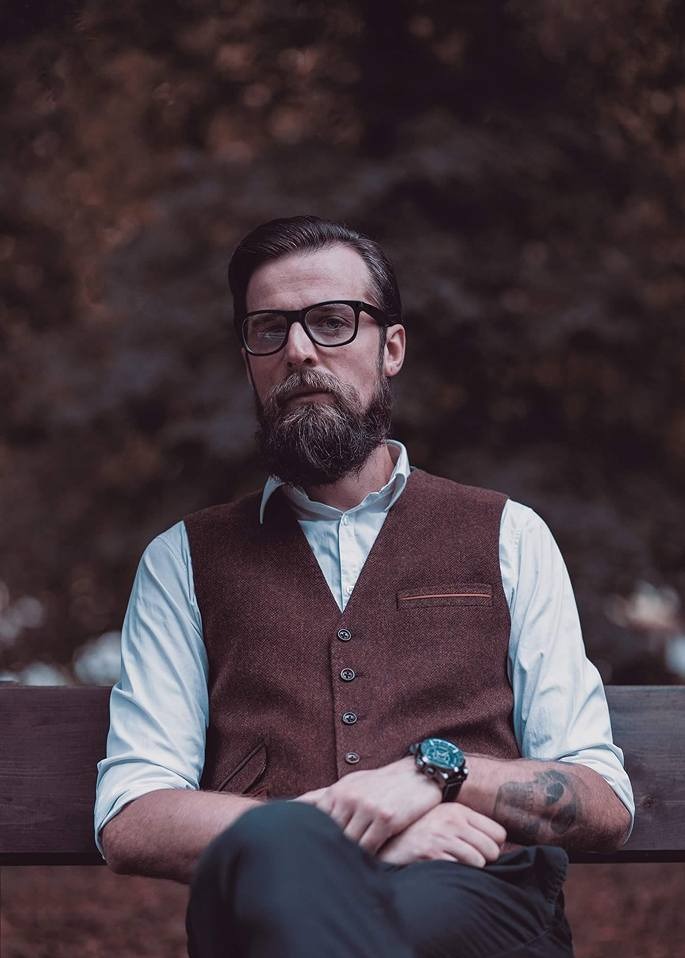 Portret brodatego mężczyzny w okularach, brązowej kamizelce i białej koszuli, siedzącego na ławce z zamyślonym wyrazem twarzy.