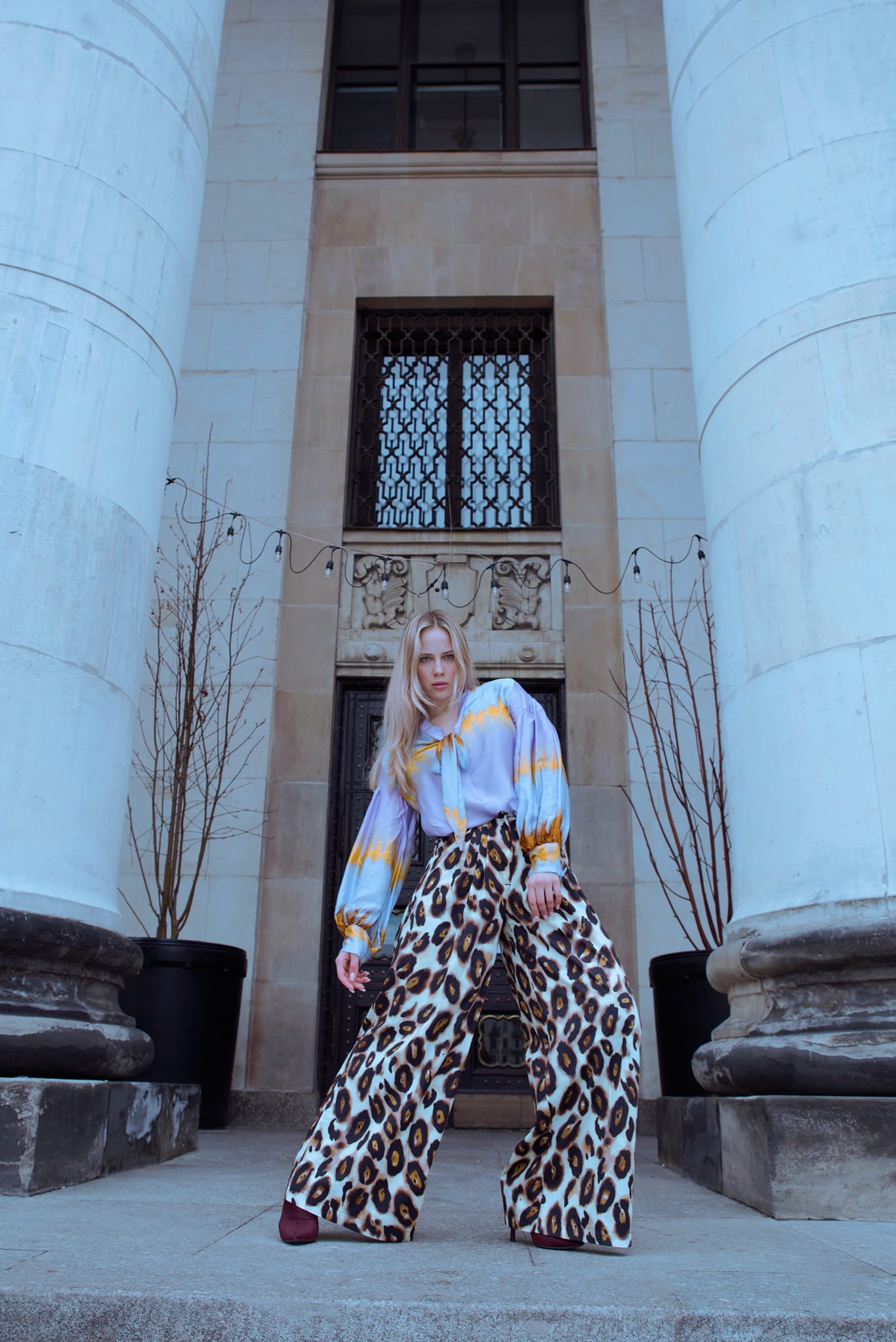 Kobieta w bluzce z bufkami i spodniach w panterkę pozuje na tle budynku z kolumnami w Warszawie. Nietypowe ujęcie z dołu podkreśla architekturę.