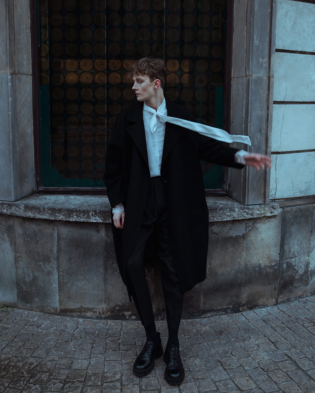 Młody mężczyzna w czarnym płaszczu i białej koszuli z rozluźnionym krawatem, pozujący na tle kamiennej ściany z okrągłym oknem i brukowanej nawierzchni.