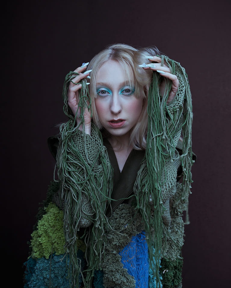 Portret kobiety z blond włosami i mocnym turkusowym makijażem oczu, ubrana w artystyczną kreację z zielonych i niebieskich włóczek oraz dzianin, pozująca na ciemnobrązowym tle.