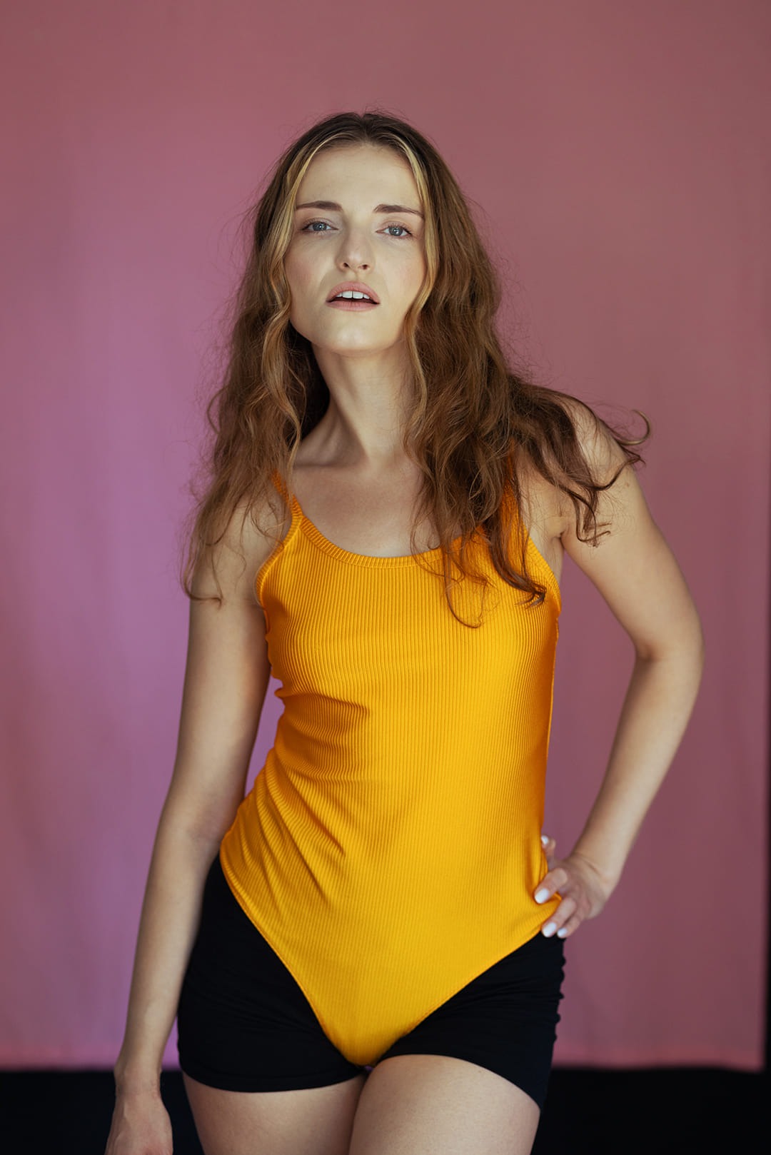 Kobieta z rozwianymi włosami pozuje w żółtym body na tle różowego studia fotograficznego.