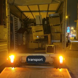 Mario Super clean - Wnętrze skrzyni ładunkowej busa wypełnione kartonami, meblami i przedmiotami podczas nocnej przeprowadzki, widoczne światła stop i podnośnik załadunkowy.