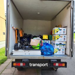 Mario Super clean - Wnętrze paki dostawczego samochodu załadowane różnorodnymi przedmiotami, w tym kartonami z bananami, workami i elementami konstrukcyjnymi, widoczne z tyłu pojazdu na tle chodnika i budynku.