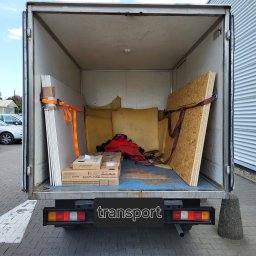 Mario Super clean - Otwarta przestrzeń ładunkowa busa wypełniona płytami gipsowymi zabezpieczonymi pomarańczowymi pasami, płytą OSB z czerwonym pasem, kartonami oraz elementami tapicerki, na zewnątrz widoczne...