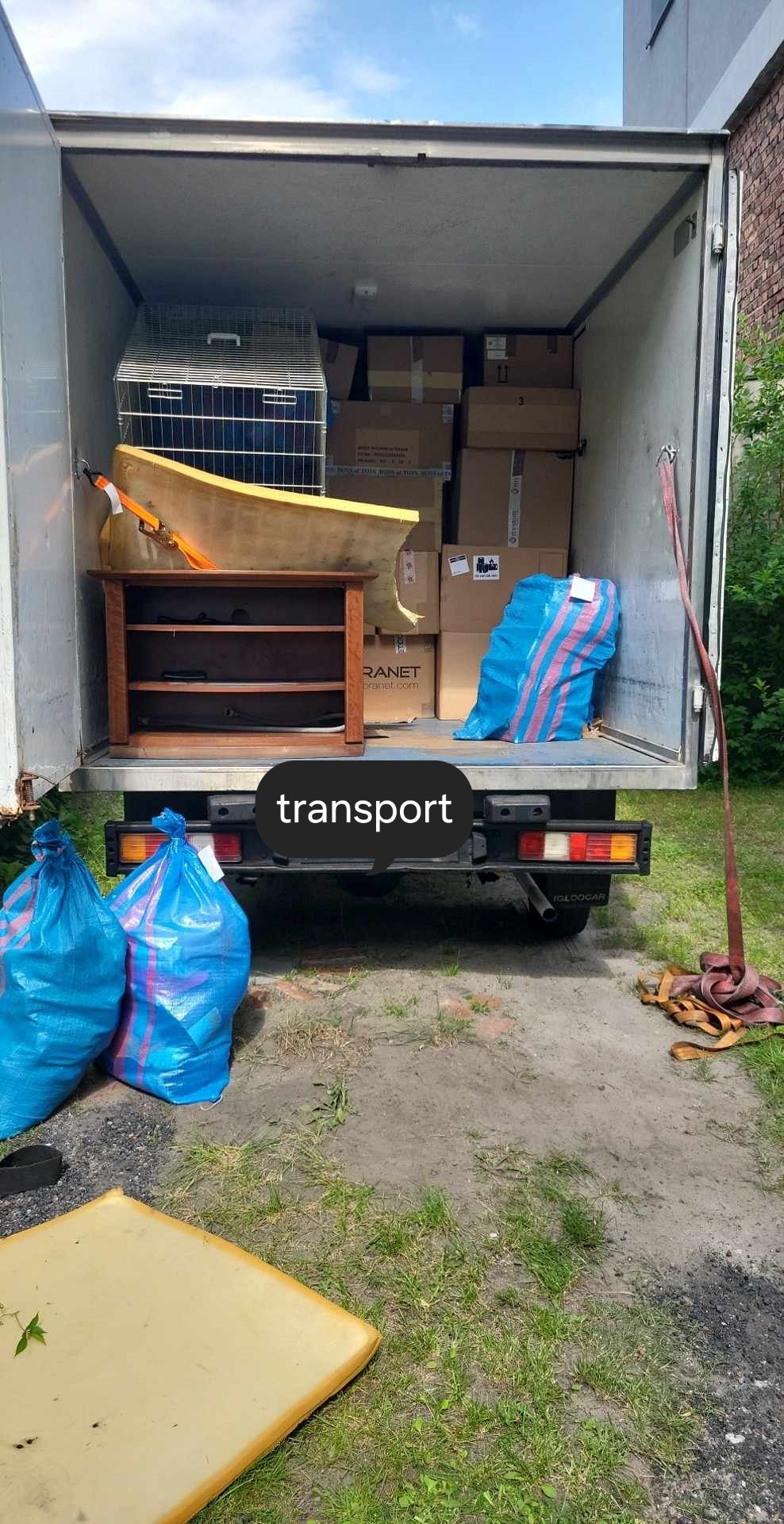 Wnętrze busa załadowane kartonami, meblami i workami, widoczne z zewnątrz, z ziemią i trawą w tle.