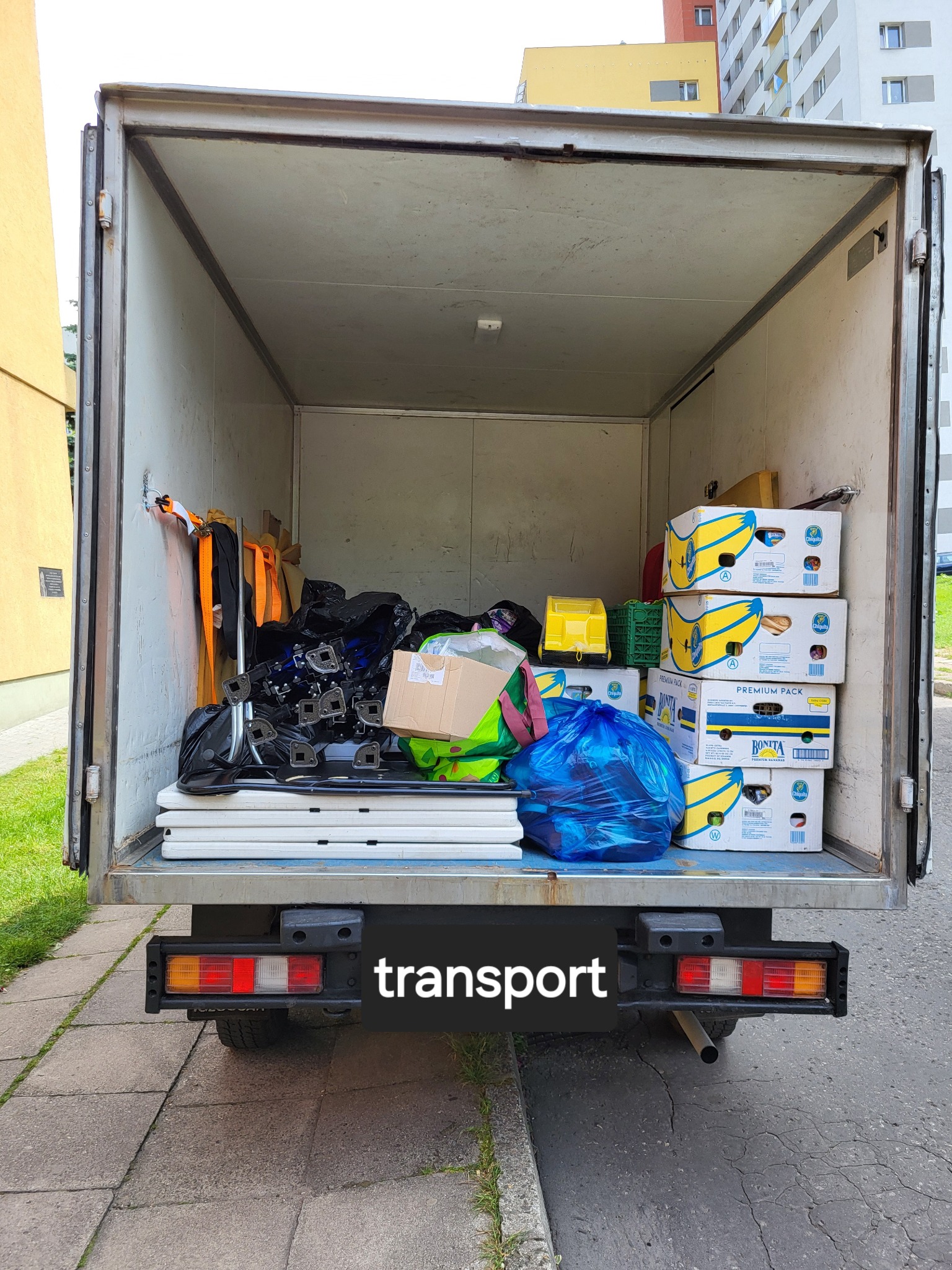 Wnętrze paki dostawczego samochodu załadowane różnorodnymi przedmiotami, w tym kartonami z bananami, workami i elementami konstrukcyjnymi, widoczne z tyłu pojazdu na tle chodnika i budynku.