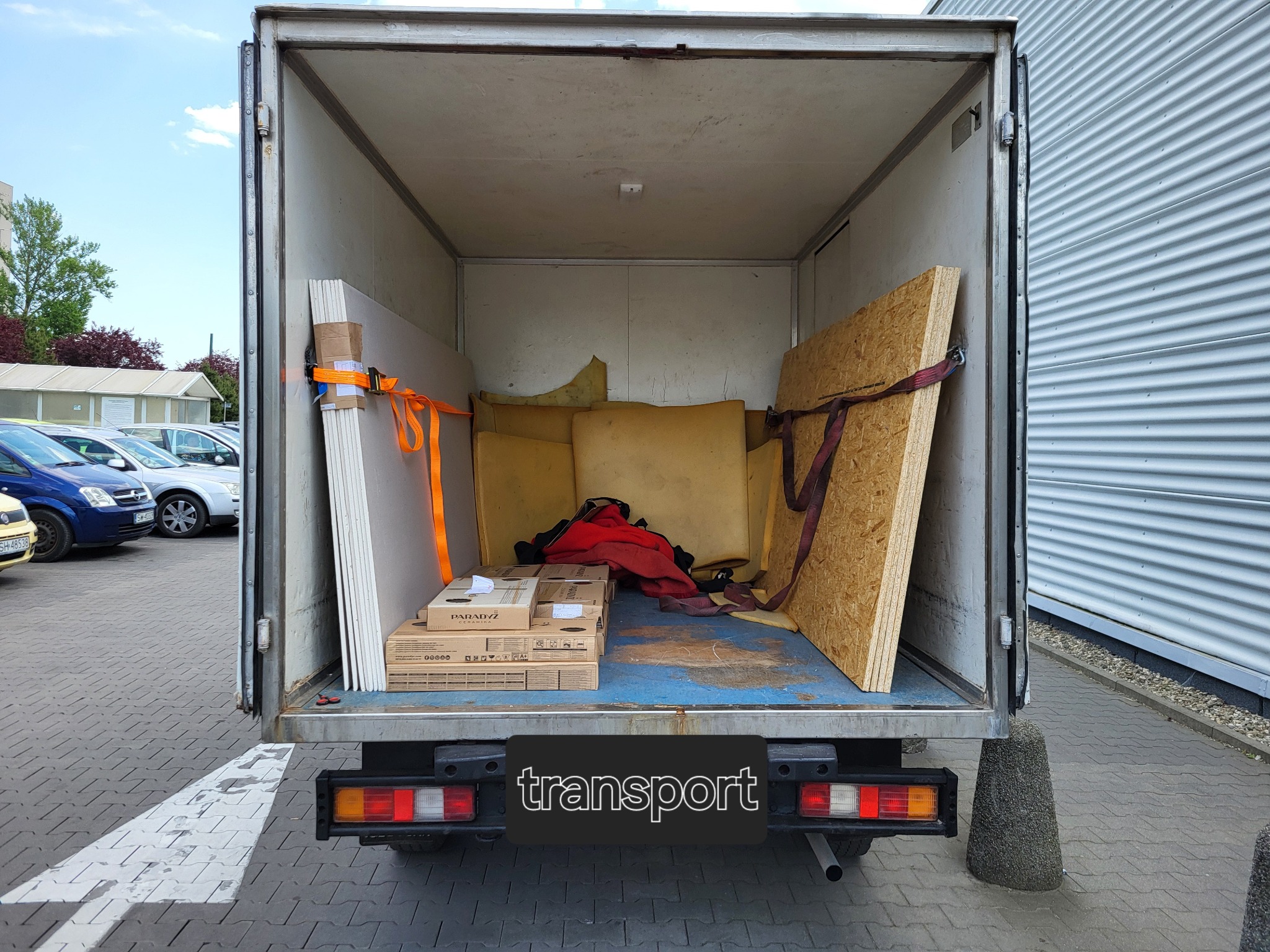 Otwarta przestrzeń ładunkowa busa wypełniona płytami gipsowymi zabezpieczonymi pomarańczowymi pasami, płytą OSB z czerwonym pasem, kartonami oraz elementami tapicerki, na zewnątrz widoczne...