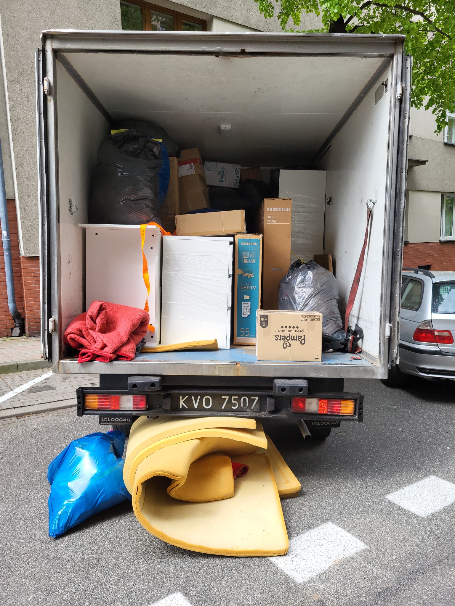 Wnętrze furgonetki załadowane meblami, kartonami (w tym z telewizorem Samsung i pieluchami Pampers) i workami na śmieci; na zewnątrz, za klapą bagażnika leżą fragmenty pianki i niebieski worek.