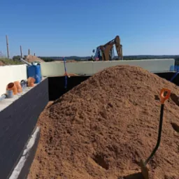 Wykop fundamentów z izolacją termiczną i hydroizolacją, widoczna góra piasku oraz rury kanalizacyjne i koparka w tle.