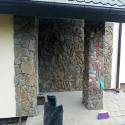 Wykończone wejście do budynku z kamienną elewacją i kolumnami. Na jednej z kolumn widoczny krzyż z kamieni. Obok leży miotła.