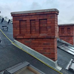 AGBS CONSTRUCTIONS SPÓŁKA Z OGRANICZONĄ ODPOWIEDZIALNOŚCIĄ - Ceglany komin z betonową czapą na czarnym dachu z oknami dachowymi. Widoczne metalowe obróbki blacharskie i żółty przewód. Pochmurne niebo w tle.