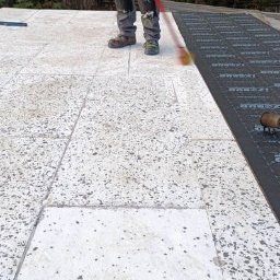 AGBS CONSTRUCTIONS SPÓŁKA Z OGRANICZONĄ ODPOWIEDZIALNOŚCIĄ - Pracownik rozprowadza masę bitumiczną na płaskim dachu za pomocą czerwonego wałka. Część dachu pokryta jest już czarną membraną izolacyjną.