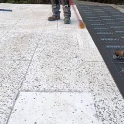 Pracownik rozprowadza masę bitumiczną na płaskim dachu za pomocą czerwonego wałka. Część dachu pokryta jest już czarną membraną izolacyjną.