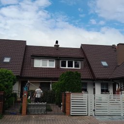 AGBS CONSTRUCTIONS SPÓŁKA Z OGRANICZONĄ ODPOWIEDZIALNOŚCIĄ - Rząd domów z brązowymi dachami z dachówki, barierki w kolorze białym. Na jednym z kominów ozdobna figurka ptaka. Osoba pracuje przy elewacji jednego z budynków.