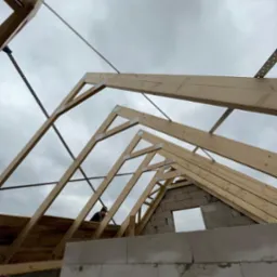 Konstrukcja drewniana dachu w trakcie budowy, widok od dołu na szkielet z belek i metalowych łączników na tle pochmurnego nieba. Mur z bloczków.
