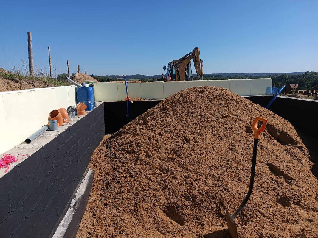 Wykop fundamentów z izolacją termiczną i hydroizolacją, widoczna góra piasku oraz rury kanalizacyjne i koparka w tle.