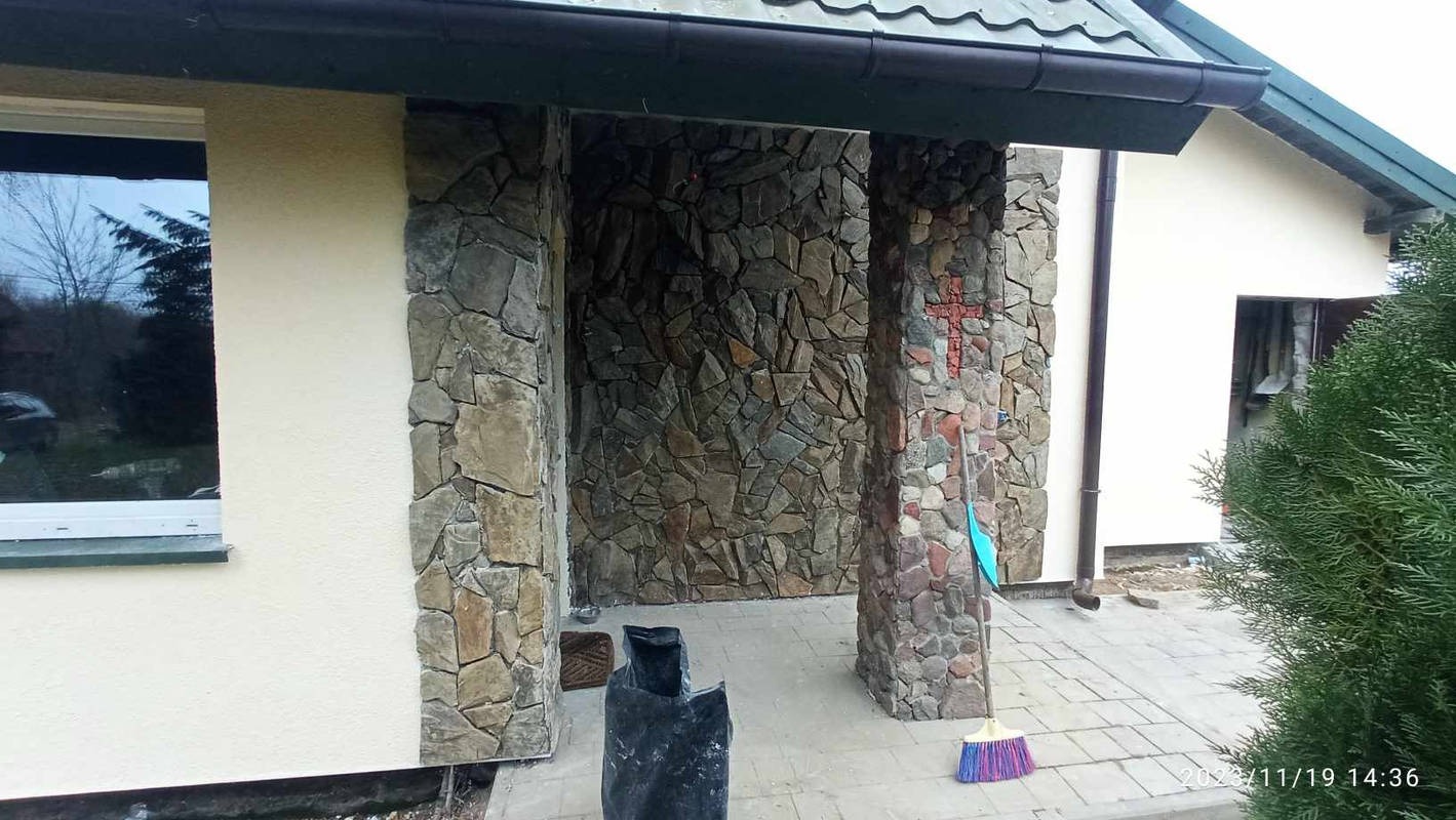 Wykończone wejście do budynku z kamienną elewacją i kolumnami. Na jednej z kolumn widoczny krzyż z kamieni. Obok leży miotła.