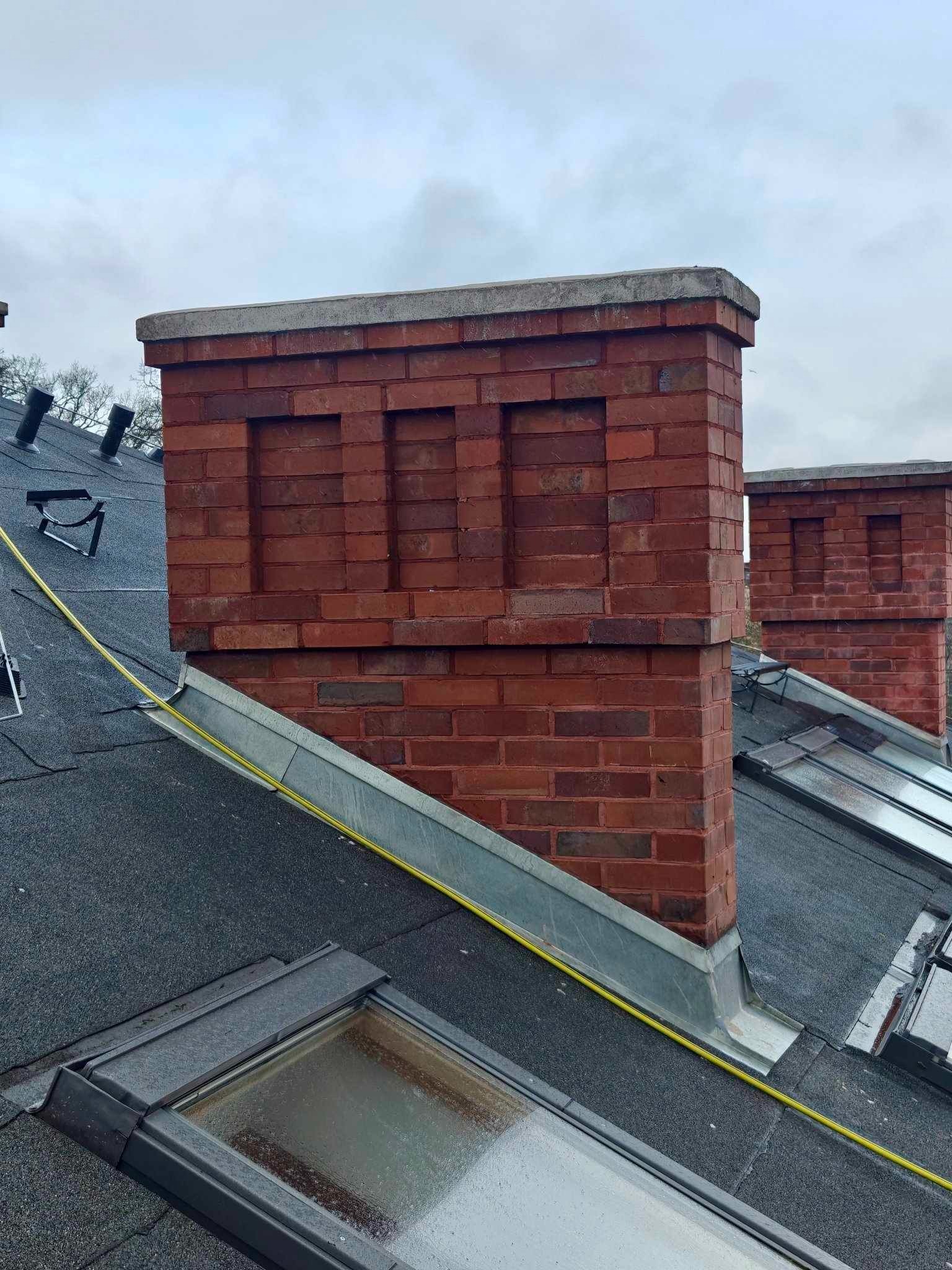 Ceglany komin z betonową czapą na czarnym dachu z oknami dachowymi. Widoczne metalowe obróbki blacharskie i żółty przewód. Pochmurne niebo w tle.