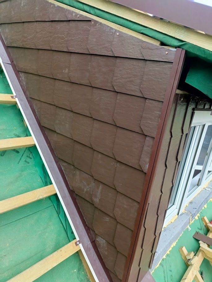 Detal montażu brązowej dachówki bitumicznej na skośnym dachu z oknem połaciowym. Widoczne elementy konstrukcyjne i membrana dachowa.